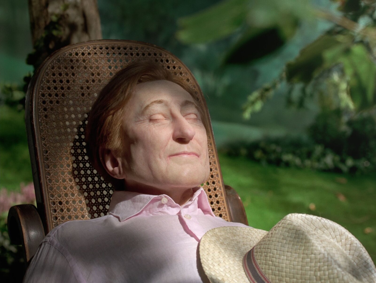 Still from La Gola by Diego Marcon: a woman with closed eyes resting in a wicker chair in a garden, a straw hat on her chest
