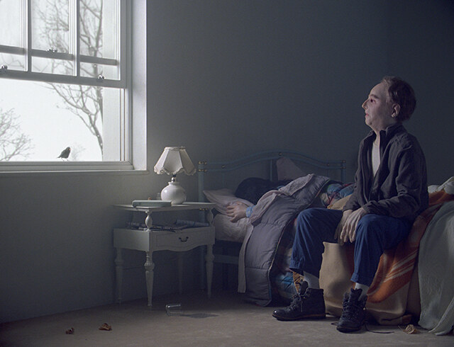 Still from The Parents' Room by Diego Marcon: a figure sitting on the edge of a bed in a dim room, looking at a blackbird on the window sill